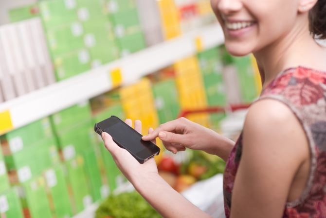 Smiling young woman at supermarket using mobile phone and looking at camera.
